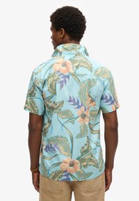Camicia a maniche corte, di colore azzurro chiaro, con un motivo floreale tropicale caratterizzato da fiori rosa e foglie verdi. Tessuto in cotone, vestibilità morbida, con colletto.