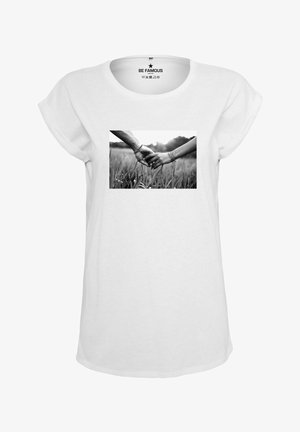 Weißes T-Shirt mit kurzen Ärmeln, das ein rechteckiges schwarz-weiß Foto zweier Hände zeigt, die sich in hohem Gras halten. Glatte Stofftextur.