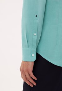Türkisfarbenes Hemd mit strukturierter Stoffqualität. Verfügt über eine geknöpfte Manschette und eine kleine schwarze Rosenstickerei nahe der Manschette. Elegantes und figurnahes Design.