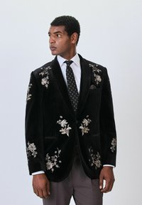 Black velvet blazer with floral embroidery in gold and silver, two front pockets, peaked lapel, worn over a white shirt and patterned tie.