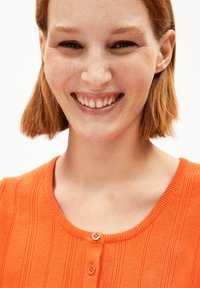 Haut orange à côtes, avec un col rond et deux boutons au centre. Le tissu a une texture lisse avec un design ajusté.