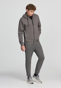 Felpa grigia con zip, tasche a marsupio e cappuccio regolabile con cordino, abbinata a pantaloni della tuta a gamba affusolata. Presenta una texture del tessuto liscia.