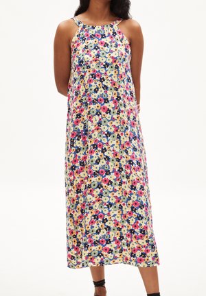Femme portant une robe sans manches, longue jusqu'aux chevilles, avec un motif floral multicolore, se tenant devant un fond blanc uni.