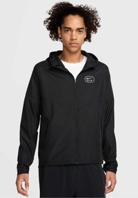 Jeune homme aux cheveux bouclés portant une veste à capuche zippée noire Nike Air sur un t-shirt blanc, debout devant un fond clair uni.