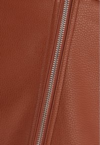 Detailní záběr na hnědou texturovanou koženou plochu se středovým stříbrným kovovým zipem probíhajícím vertikálně.