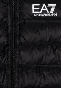 Gilet piumino nero con chiusura a zip, caratterizzato da sezioni trapuntate orizzontali e un logo EA7 Emporio Armani bianco sul petto.