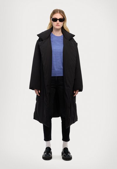 Black oversized puffer coat with large collar, worn over a blue sweater. Black pants, white socks, and chunky black shoes complete the look.