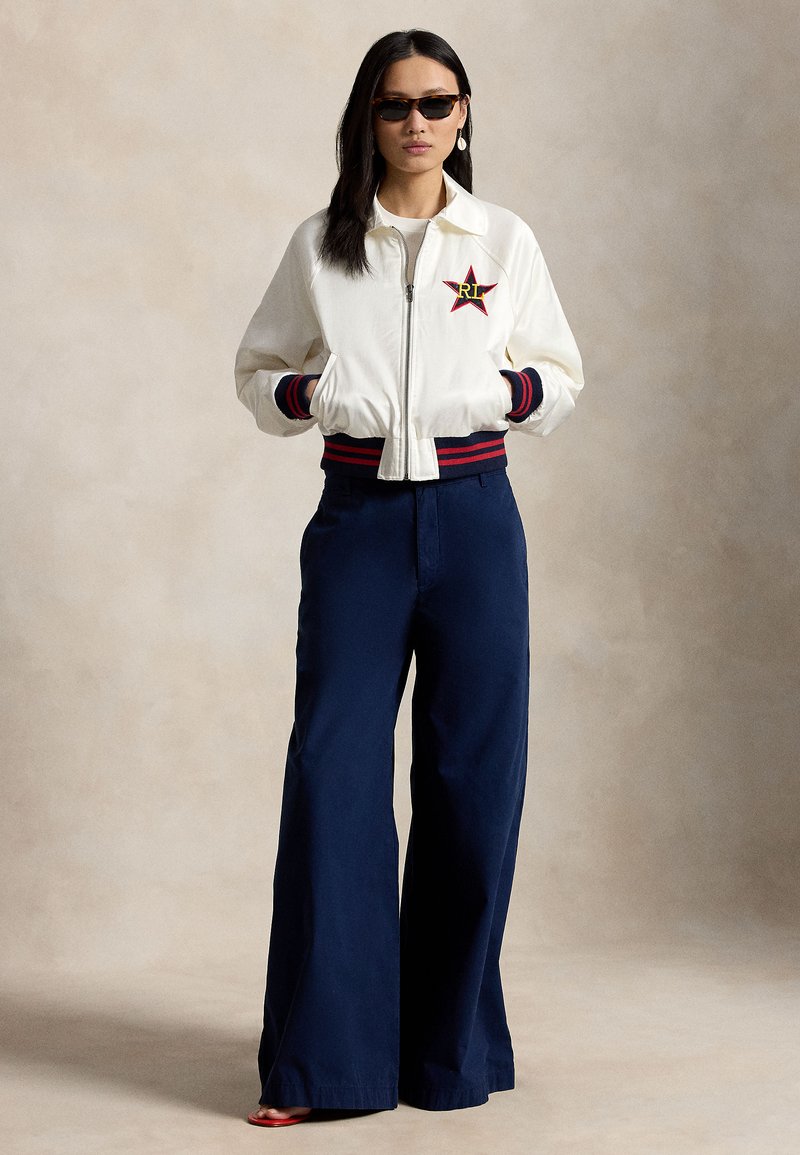 Femme portant une veste bomber blanche avec un emblème en forme d'étoile, un pantalon large bleu marine, des lunettes de soleil et des sandales rouges, debout devant un fond neutre.