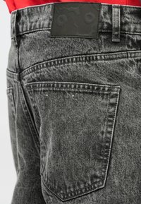 Jeans de ganga cinza com acabamento texturizado, bolsos traseiros padrão e um remendo de couro preto no cós.