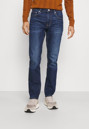 Person, die slim-fit dunkelblaue Jeans, hellbeige Sneakers und einen orangenen Langarmpullover trägt, steht vor einem schlichten hellen Hintergrund.