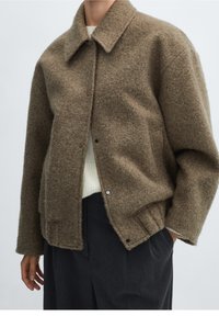 Brown textured wool jacket with a collared neckline, front snap closure, and two side pockets, worn over a light sweater and dark trousers.