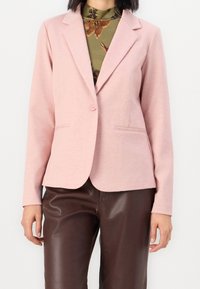 Blazer rosa claro fabricado en tela suave, con cierre de un solo botón, solapas de muesca y dos bolsillos frontales. Combinado con pantalones de cuero sintético marrones.