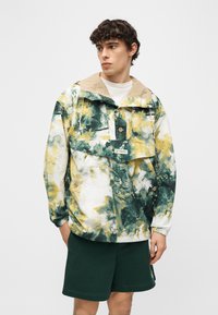 Νεαρός άνδρας που φοράει φούτερ με κουκούλα σε αποχρώσεις πράσινου, κίτρινου και λευκού με σχέδιο tie-dye και σκούρα πράσινα σορτς, στέκεται με το χέρι στην τσέπη κοιτώντας αριστερά.