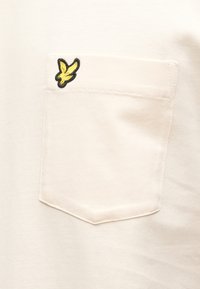 Lys cremefarvet t-shirt med korte ærmer, der har en brystlomme på venstre side med et gult og sort broderet fugledesign. Blød bomuldstekstur.