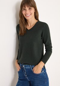 Dunkelgrüner V-Ausschnitt-Pullover mit langen Ärmeln, figurbetontem Design, weicher Textur. Getragen mit blauen Jeans mit Knopfverschluss und Eingrifftaschen.
