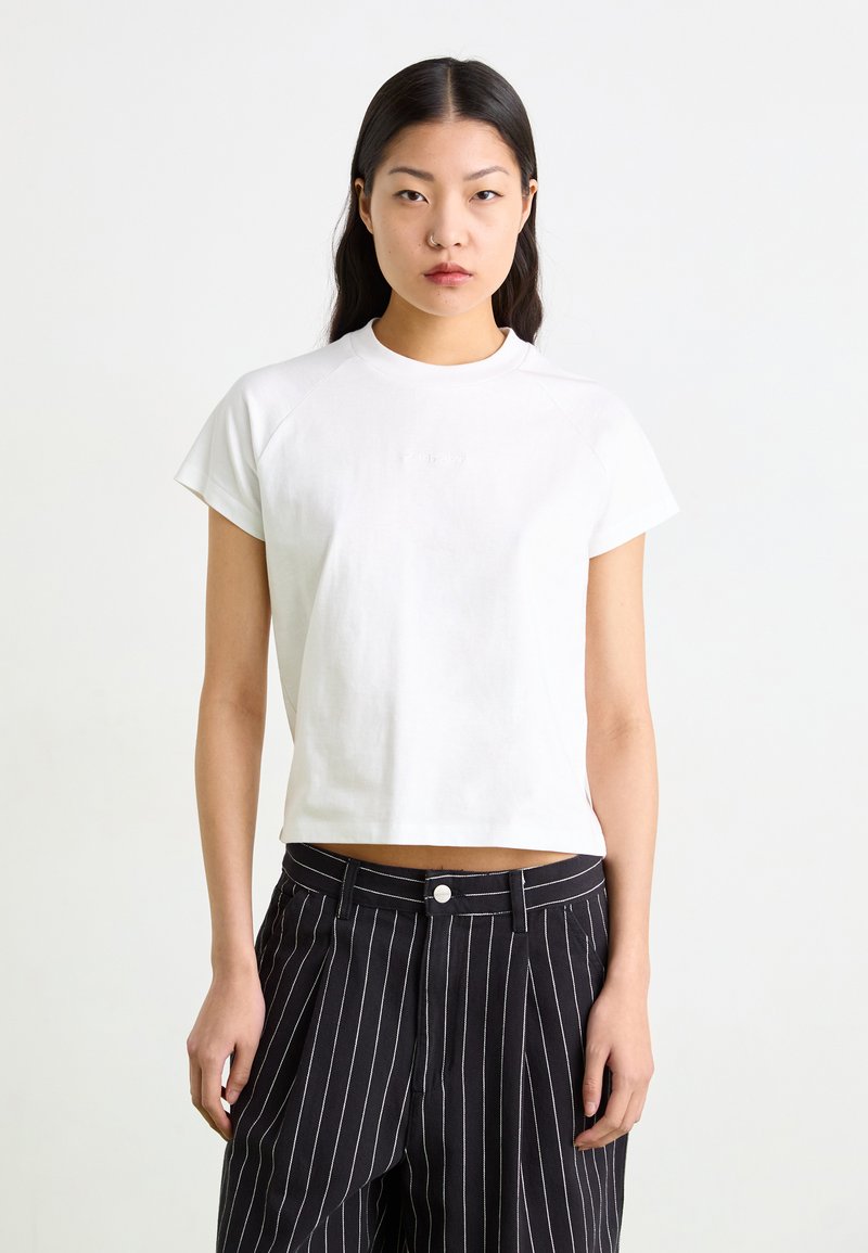 Hvid cropped t-shirt med korte raglanærmer, rund halsudskæring og subtilt præget logo. Sammen med sorte bukser med smalle striber.