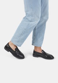 Mocasines de cuero negro con diseño de punta cosida y un tacón bajo, combinados con jeans de tiro alto y puños azul claro, mostrando un look casual.