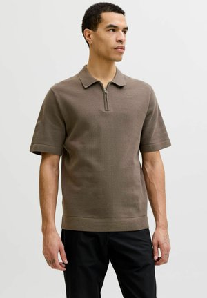 Uomo che indossa una polo marrone a maniche corte con zip e pantaloni neri, in piedi davanti a uno sfondo chiaro e uniforme.