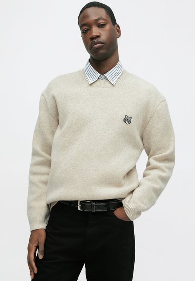 Bēžs džemperis ar garām piedurknēm un ribotu apkakli, ar izšūtu pelēku vilku galvu. Kombinēts ar svītrainu krekli un melnām biksēm.