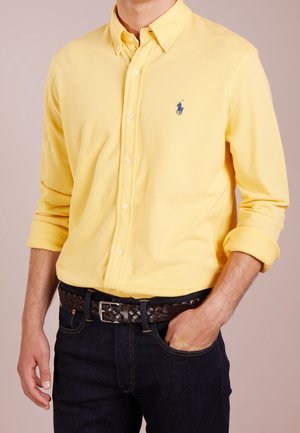 Homme portant une chemise à manches longues jaune avec un logo bleu brodé, un jean foncé et une ceinture en cuir brun tressé.