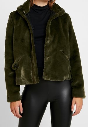 Vrouw draagt een donkergroene jas van nepbont over een zwarte coltrui en zwarte leren broek, staand tegen een effen achtergrond.