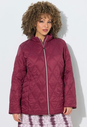 Donna con capelli ricci che indossa una giacca trapuntata color bordeaux con cerniera frontale e gonna con motivo, in piedi su uno sfondo chiaro uniforme.