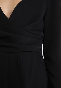 Vestido negro con escote en V profundo y mangas largas. Confeccionado en tela suave, presenta un diseño cruzado en la cintura y una falda fluida.