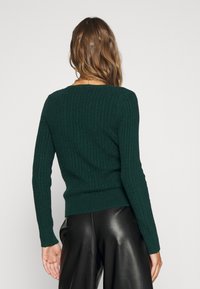 Suéter verde con canalé, de manga larga y cuello redondo, hecho de una tela de punto texturizada, combinado con pantalones de cuero negros.