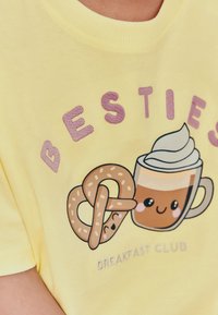 Gul bomulls t-shirt med en söt grafik av en kaffekopp och en pretzel, med "BESTIES" i lila broderi och "BREAKFAST CLUB" nedanför.