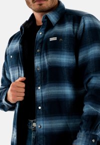 Heren flanellen overhemd in donkerblauw met lichtblauwe strepen, drukknoopsluitingen, twee borstzakken en een label op de linker zak. Zachte textuur.