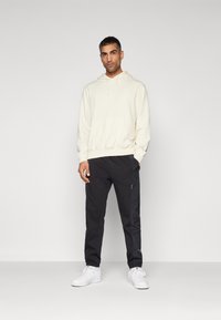 Cream hoodie with front pocket, paired with black jogger pants featuring side pockets and white sneakers. Textured fabric and fitted design.