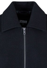 Jachetă neagră cu fermoar și guler larg, cu fermoar argintiu și etichetă "Urban Classics XS" în interiorul gulerului.