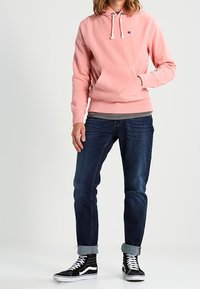 Roze hoodie met een kangoeroezak, capuchon met trekkoord en geborduurd logo. Gecombineerd met donkerblauwe spijkerbroek en zwarte hoge sneakers.