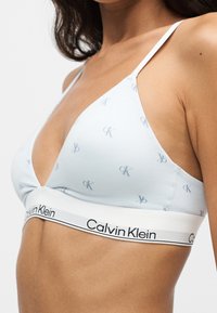 Γυναίκα που φοράει λευκό τριγωνικό σουτιέν Calvin Klein με μικρά γκρι λογότυπα CK και ελαστική ταινία με λογότυπο κάτω από το στήθος.