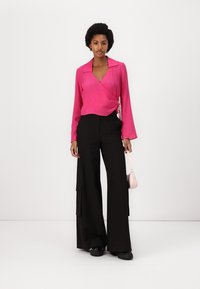 Pink wrap blouse with a collar, paired with wide black trousers featuring side pockets. The outfit is completed with black footwear and a small pink bag.