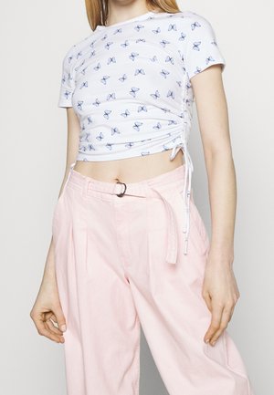 Frau trägt ein weißes, kurzes T-Shirt mit blauen Schmetterlingen und hochtaillierte rosa Hose mit Stoffgürtel und Metallschnalle.