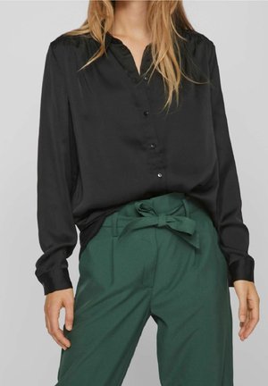 Femme portant un chemisier noir boutonné, rentré dans un pantalon taille haute vert foncé avec une ceinture en tissu nouée en nœud à la taille.