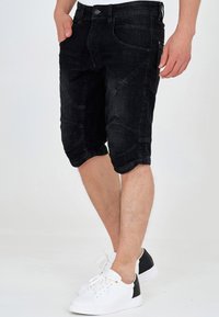 Sorte denimshorts med tætsiddende design, knælangt snit, synlige sømme og en lomme foran, kombineret med hvide sneakers.