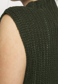 Pletený top bez rukávů v tmavě zelené barvě, s texturovanou látkou s žebrovaným vzorem a vysokým límcem. Detailní záběr na oblast ramene.