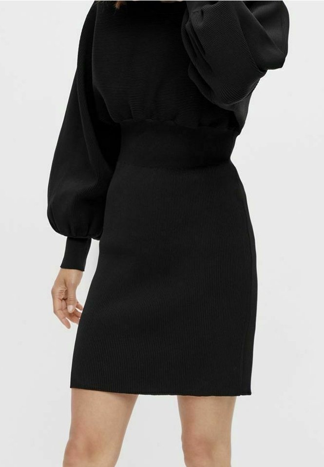 Femme portant une robe noire moulante en maille côtelée avec des manches longues bouffantes et une taille haute, debout devant un fond clair uni.
