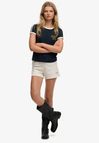 Marineblauw T-shirt met witte accenten en gele "SDR ATHL" opdruk, gecombineerd met beige korte broek en zwarte kniehoge laarzen, met gekruiste armen staand.