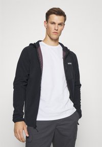 Svart zip-up hettegenser med mønstret innvendig fôr, har en frontlomme og justerbar snor i hetta. Brukt over en hvit t-skjorte.
