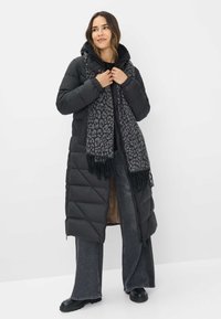 Vrouw in een lange zwarte pufferjas, met een sjaal in luipaardprint, donkere wijde jeans en zwarte laarzen, staande tegen een witte achtergrond.