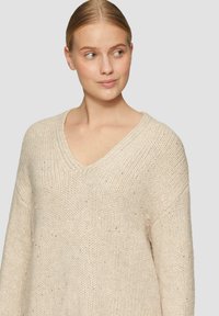 Beige gebreidetrui met V-hals, ruime pasvorm en gespikkelde metallic accenten. Zachte textuur en brede mouwen verbeteren het comfort.