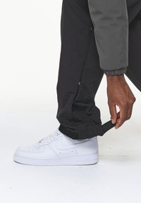 Černé kalhoty s texturovaným povrchem a elastickými manžetami, kombinované s bílými teniskami Nike Air Force 1. Osoba si upravuje manžetu pomocí pásku.