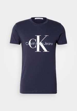T-shirt de algodão azul marinho com mangas curtas, apresentando um grande logo branco "CK" e o texto "Calvin Klein Jeans". Textura suave, corte clássico.