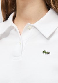 Gros plan d'une personne portant un polo blanc à col avec un petit logo crocodile vert sur la poitrine.