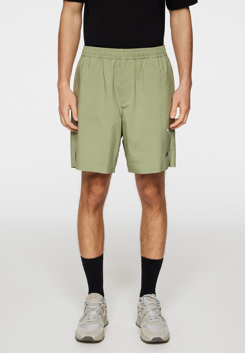 Vihreät lyhyet shortsit, joissa on joustava vyötärö, sivutaskut ja logo vasemmalla jalassa. Yhdistetty harmaisiin lenkkitossuihin ja mustiin sukkiin.