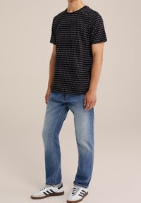 Man in een zwart gestructureerd korthoek T-shirt, lichterblauwe spijkerbroek en witte sneakers met zwarte strepen, staande tegen een beige achtergrond.