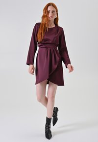 Abito di satin bordeaux con maniche lunghe, arricciatura in vita e orlo asimmetrico drappeggiato. Abbinato a stivaletti neri a punta con borchie.